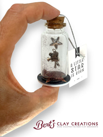 Hand holding a small glass bottle with star-shaped object and cork lid, branded 'Bert's Clay Creations'.